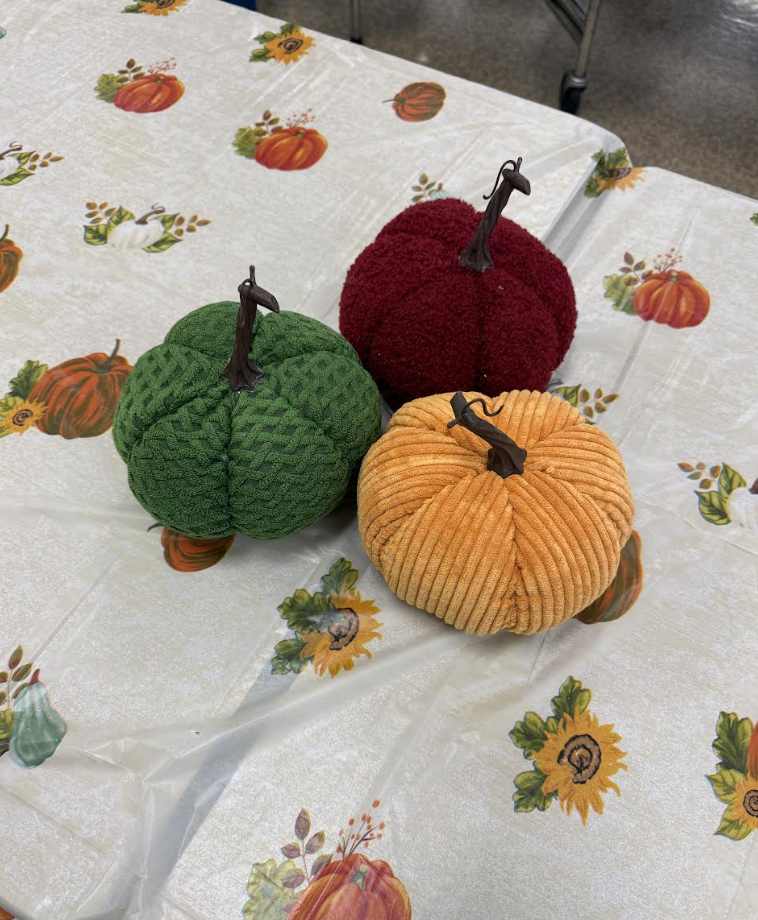 Soft, fabric pumpkins in green, burgundy, and orange sit on a fall-themed tablecloth decorated with pumpkins and sunflowers.