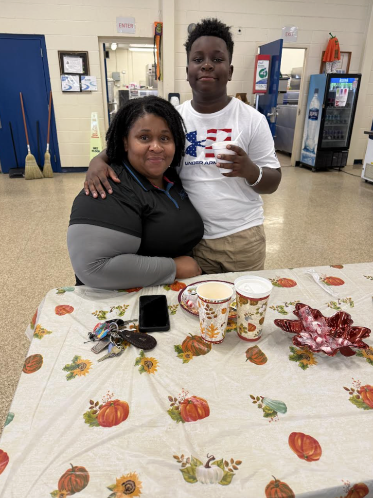 A woman sits at a decorated table with a young boy standing beside her, both looking at the camera inside a community room.