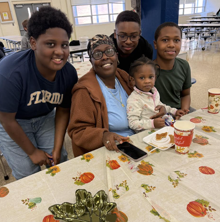 A woman sits at a decorated table with four children standing around her, in a cafeteria setting. Food and drinks are on the table.