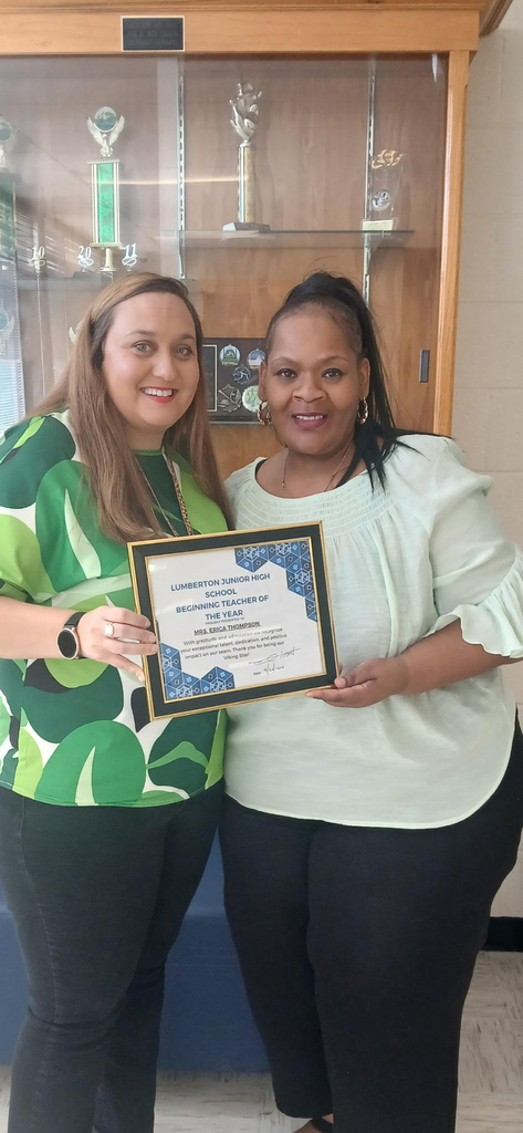 Two women stand side by side in front of a glass trophy case, smiling at the camera while holding a framed certificate. The certificate reads “Lumberton Junior High School Beginning Teacher of the Year” and is awarded to Mrs. Erica Thompson. One woman wears a green patterned blouse and a smartwatch, and the other wears a light green top with her hair pulled back. Behind them, trophies and awards are displayed on shelves, suggesting a school setting.
