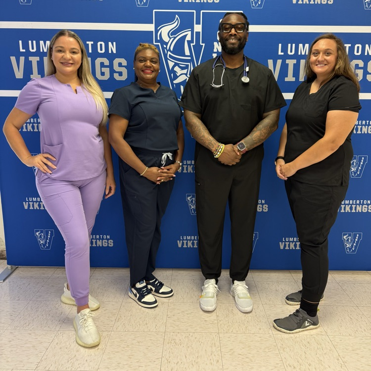 Four school administrators pose together in front of a blue-and-white Lumberton Vikings backdrop. They are wearing purple, navy, and black medical scrubs, with one person wearing a stethoscope. All four are smiling and facing the camera.