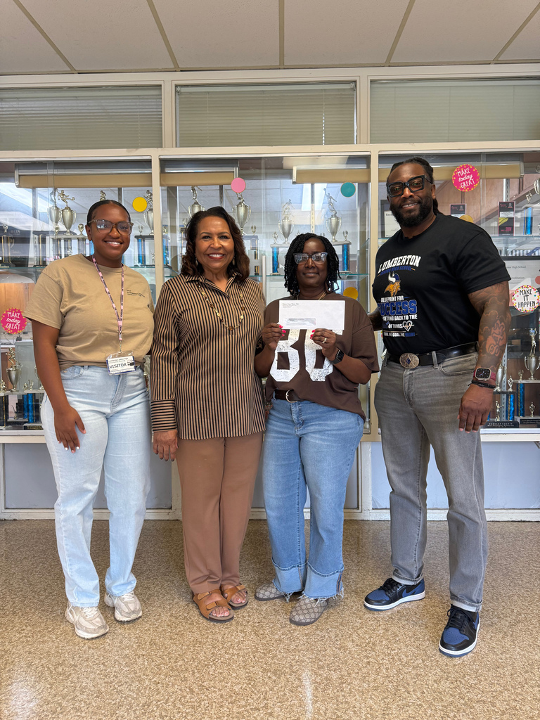 Four adults stand together smiling in front of a school trophy case. The woman near the center holds an envelope/check while the group poses for a recognition photo.