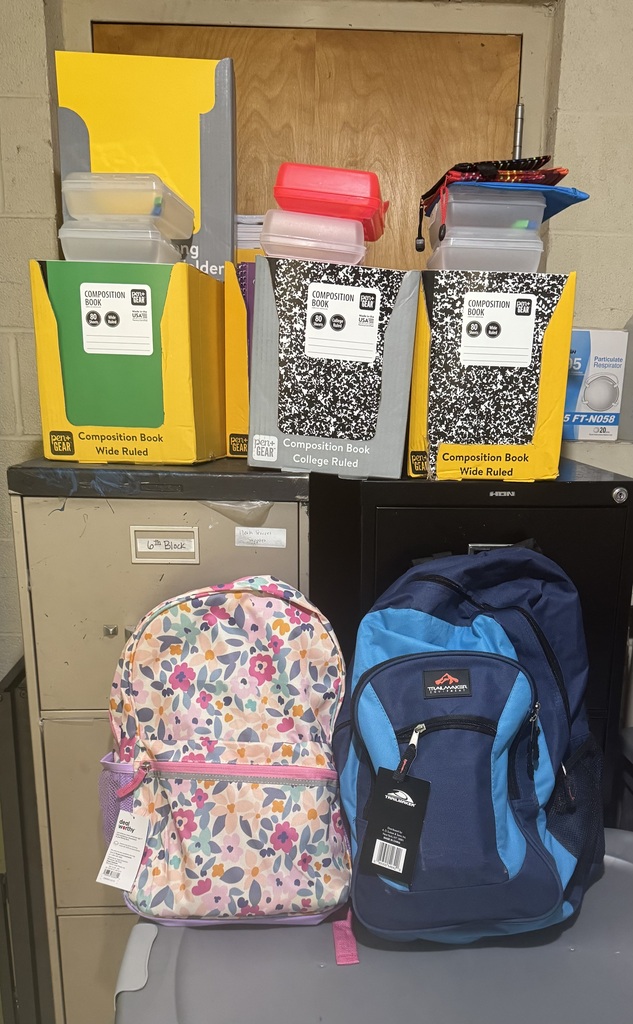 Two new backpacks, one floral and one blue, sit in front of filing cabinets. Behind them are boxes of composition notebooks, pencil boxes, folders, and other school supplies.