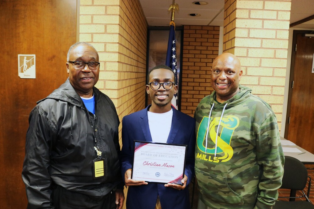 a student standing between two teachers. He is holding an award that says Christian Mason October Student Board of Education Member