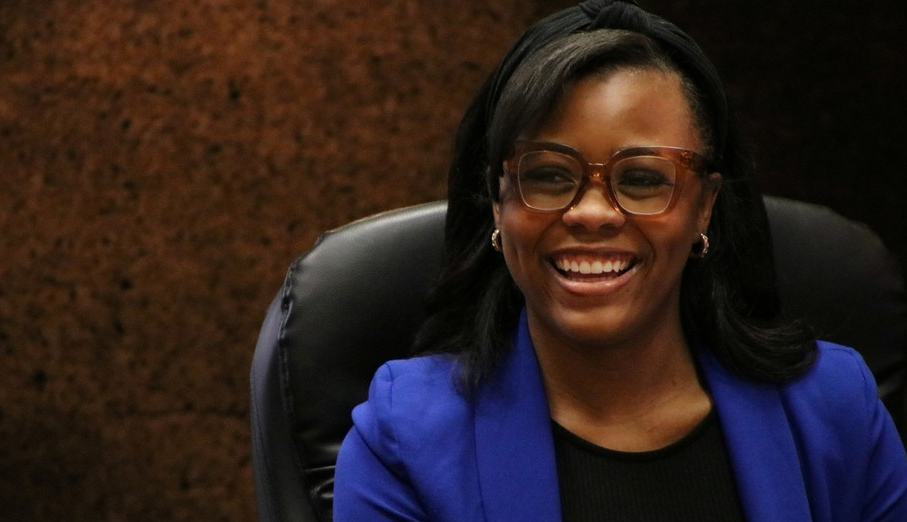 Girl smiling in glasses and blue blazer. She is sitting and has a sign that says "STUDENT" in front of her.