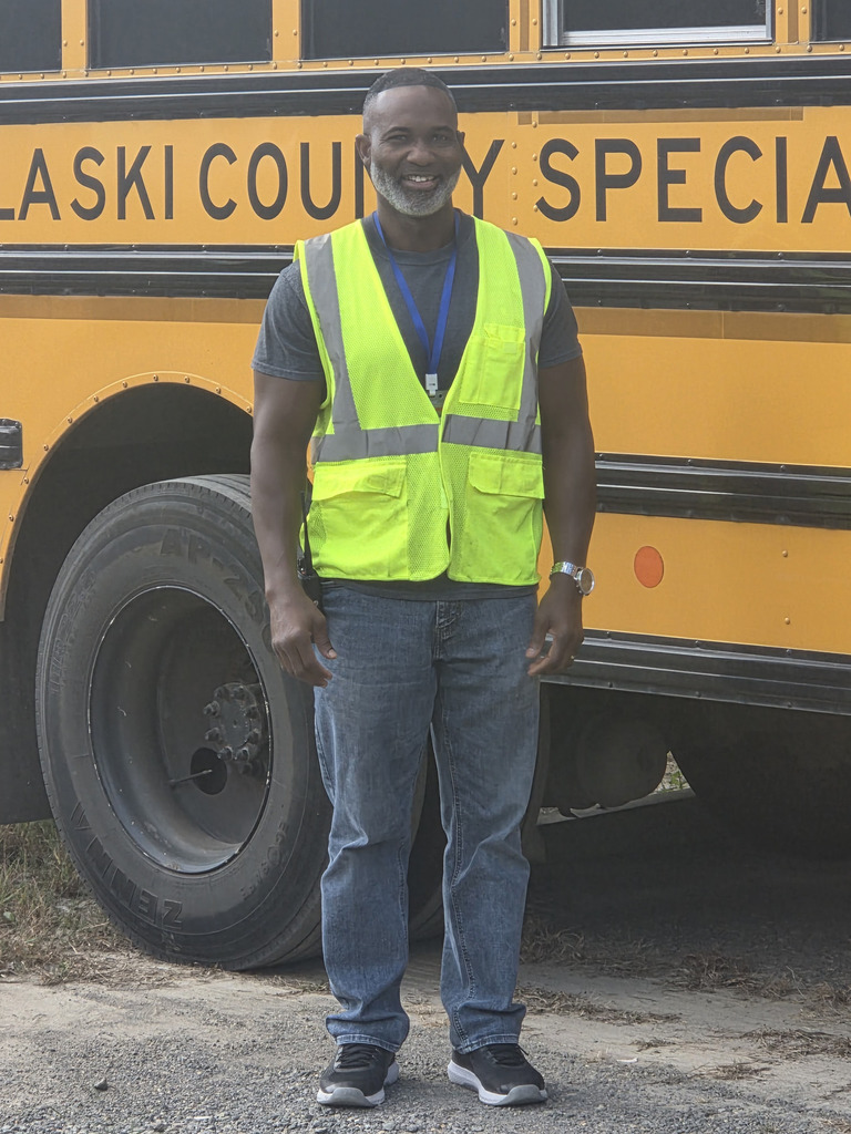 Charles Anderson standing in front of Pulaski County Special School District bus. The tall, muscular man is wearing a safety vest, lanyard, and watch.