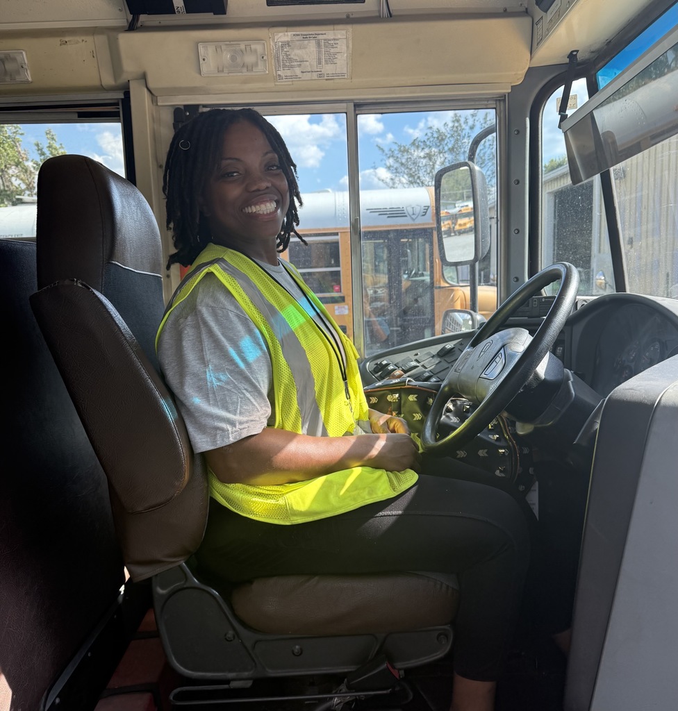 Temika Parker smiling  wearing a bright yellow safety vest and sitting in the driver's seat of a school bus.