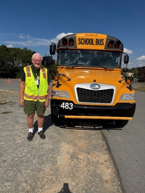 A person in a yellow safety vest stands in front of a large yellow school bus with the text "School Bus" displayed prominently.