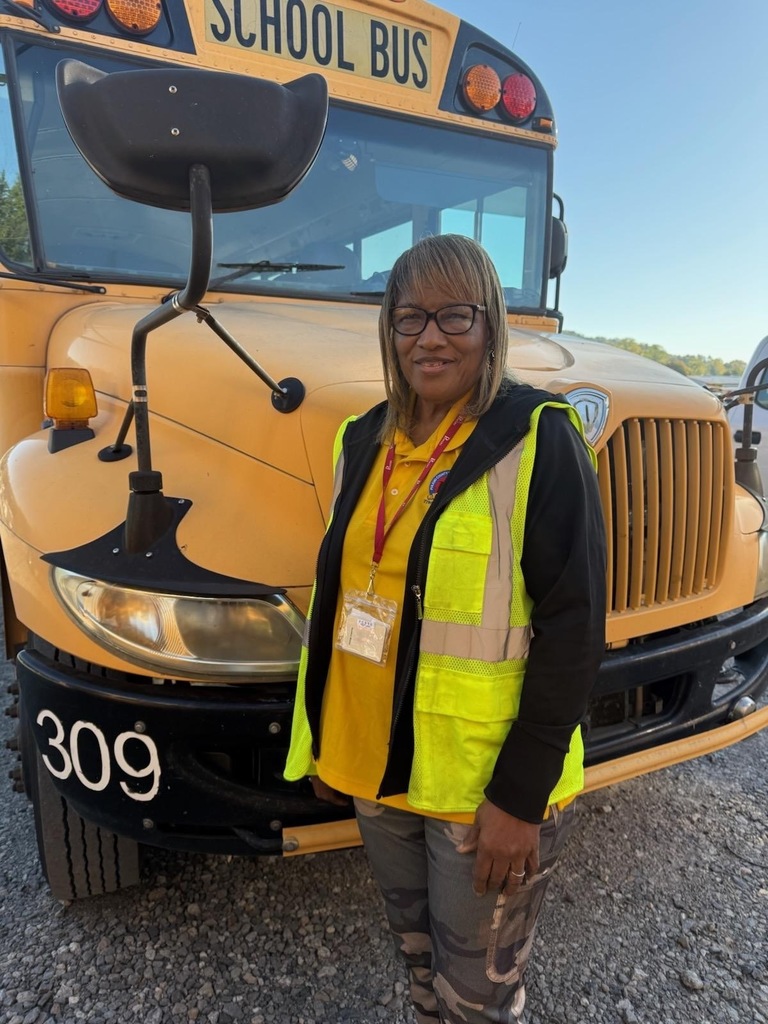 Bernice Hooks, a smiling woman in a yellow high-visibility vest and glasses standing in front of a large yellow school bus with the number "309" displayed.