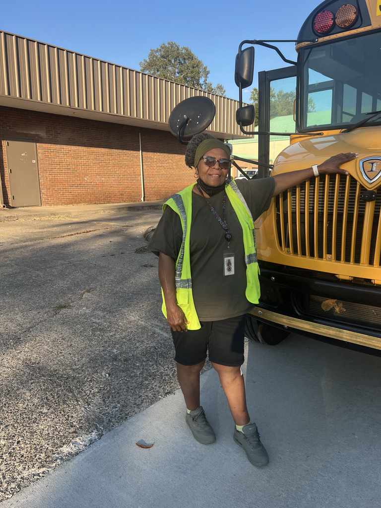 Lila White -Robinson  in a yellow safety vest and soft headband standing next to a large school bus in a parking lot.