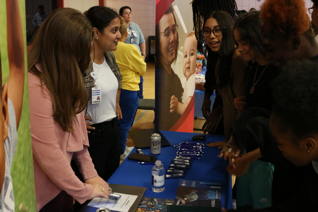 A group of people, including several young women, gathered at a table. There are papers, pens, and other materials on the table, suggesting a meeting or workshop.