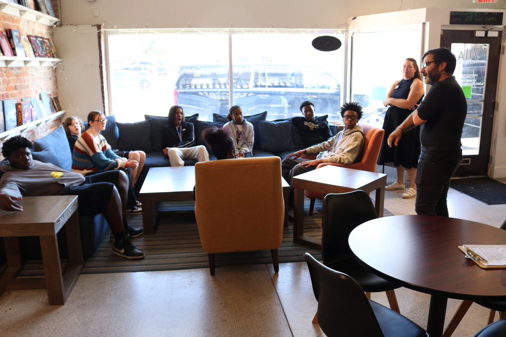A group of people sitting in a cozy cafe, some with laptops and books, engaged in conversation or working.