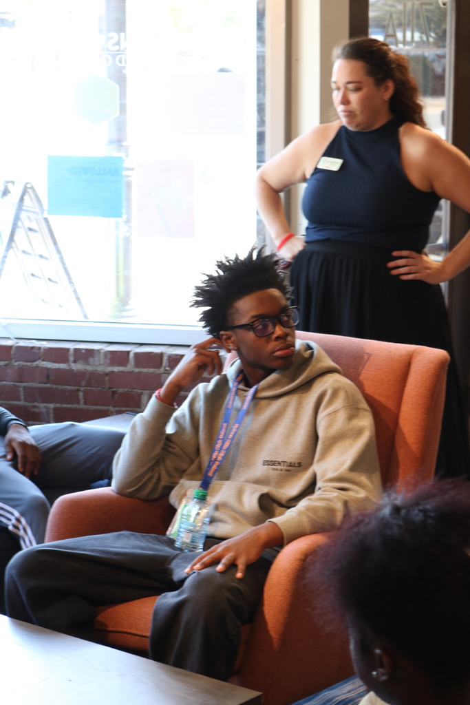 In a cozy setting, a person wearing a hooded sweatshirt sits on a couch while another person stands nearby.