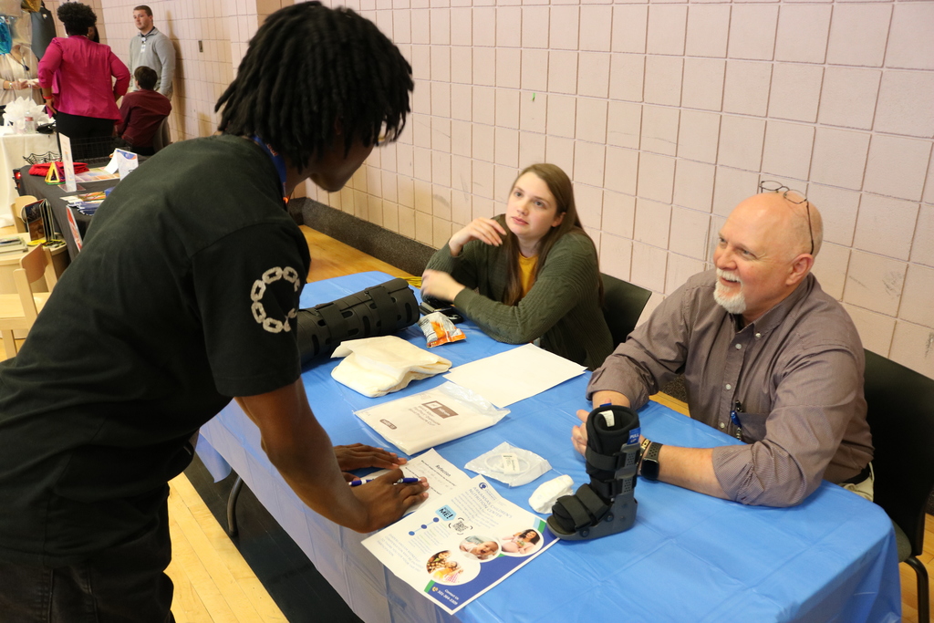A group of people sitting at a table, including a young man, a middle-aged woman, and an older man. They appear to be in discussion about casts to help people with healing.