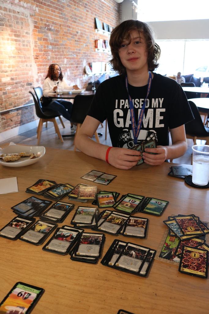 A young person in a black t-shirt with the text "I paused my game" sitting at a table surrounded by playing cards and gaming-related items.