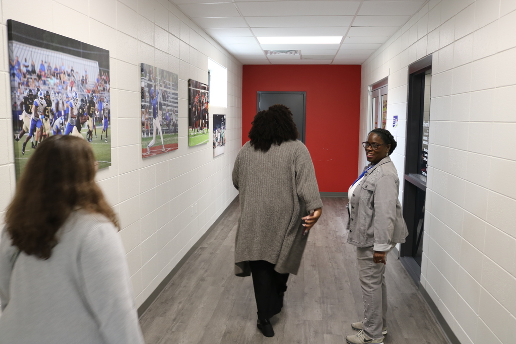 The image shows a hallway with a red accent wall and several framed photos on the walls. Three women walking down the hallway.