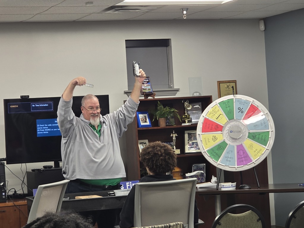 A man in a green shirt standing in front of a large colorful wheel, gesturing with his arms raised, in what appears to be a classroom or office setting.
