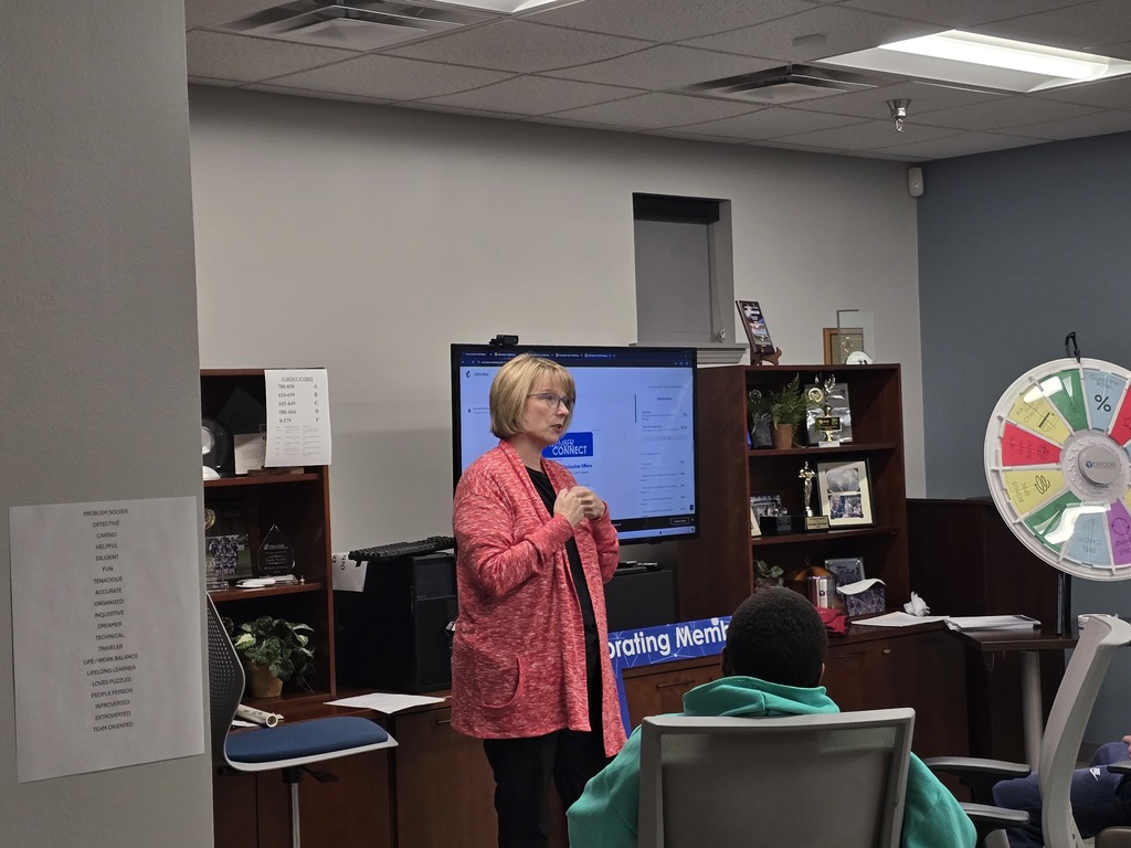 A woman in a pink cardigan speaking into a microphone while standing in front of a large screen displaying information.
