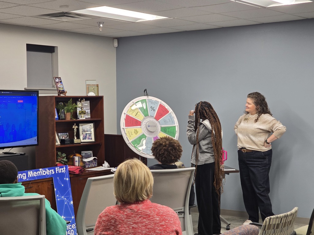 A small group of people gathered in an office setting, with a large TV screen and a wheel-like game display visible.
