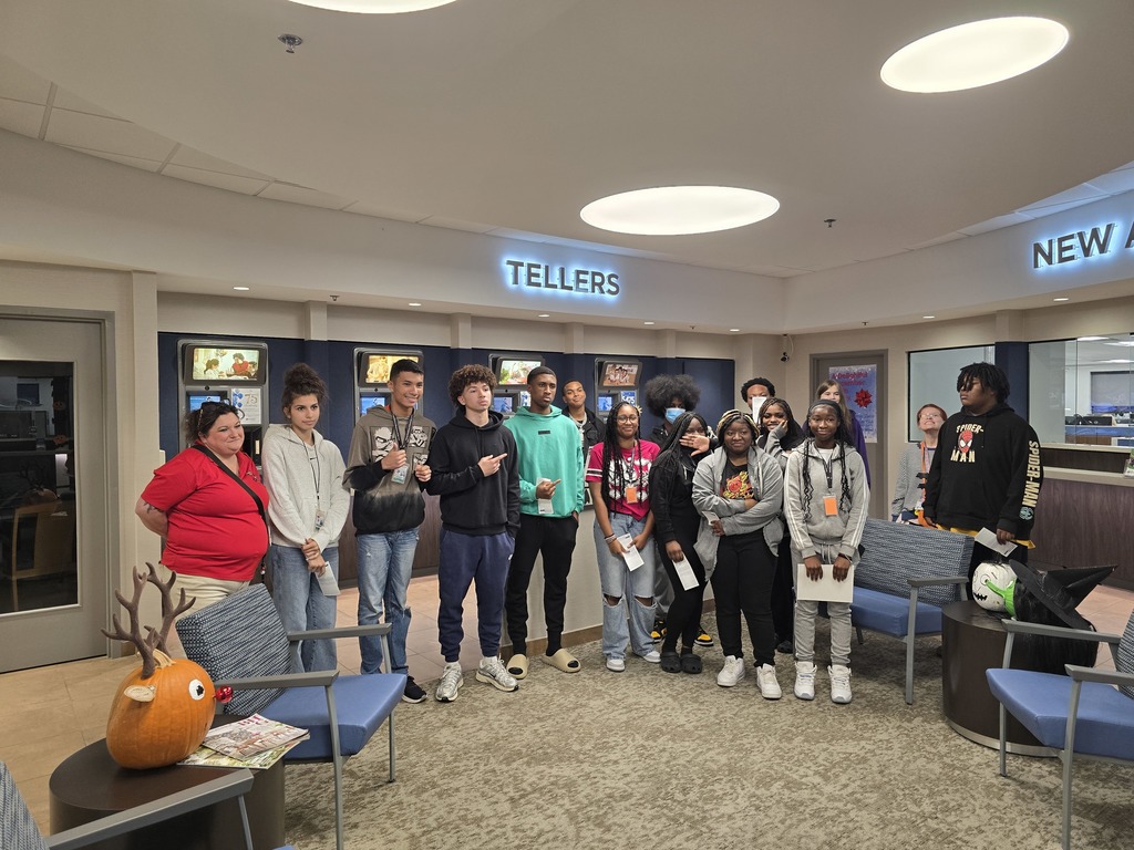 A group of people standing together in what appears to be a bank lobby or financial institution. Some are wearing casual attire, others in business casual or formal wear. Tellers and New Accounts signage visible.