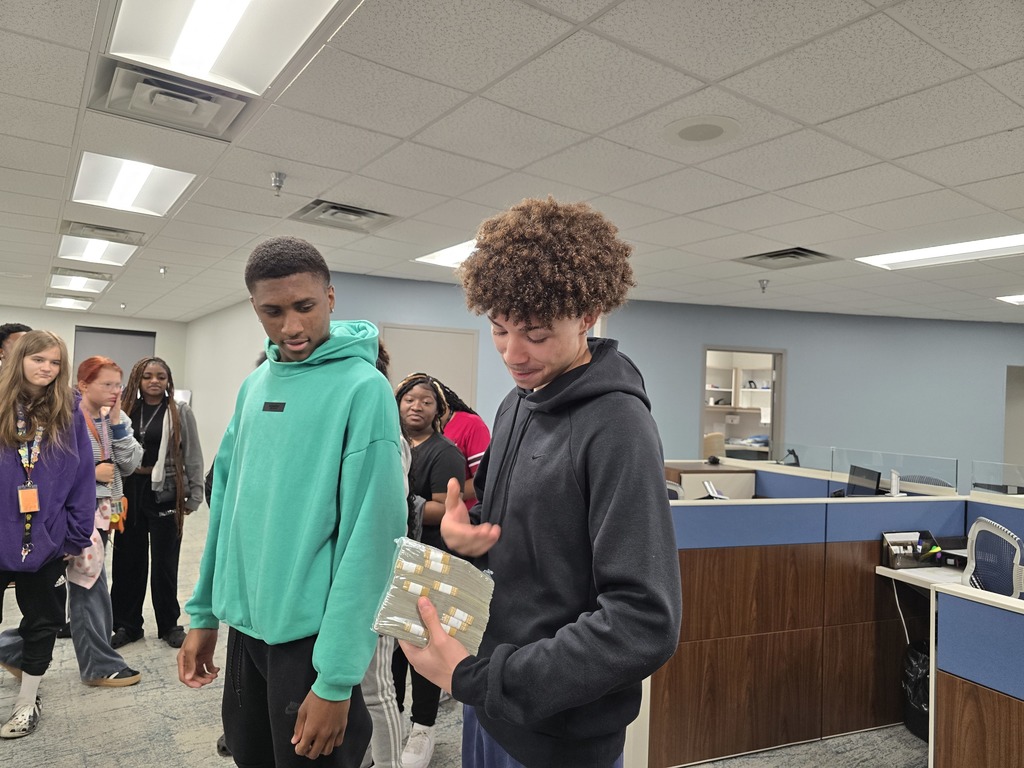 Three young people standing in an indoor setting, two wearing sweatshirts and one holding $100,000 in 100 bills.