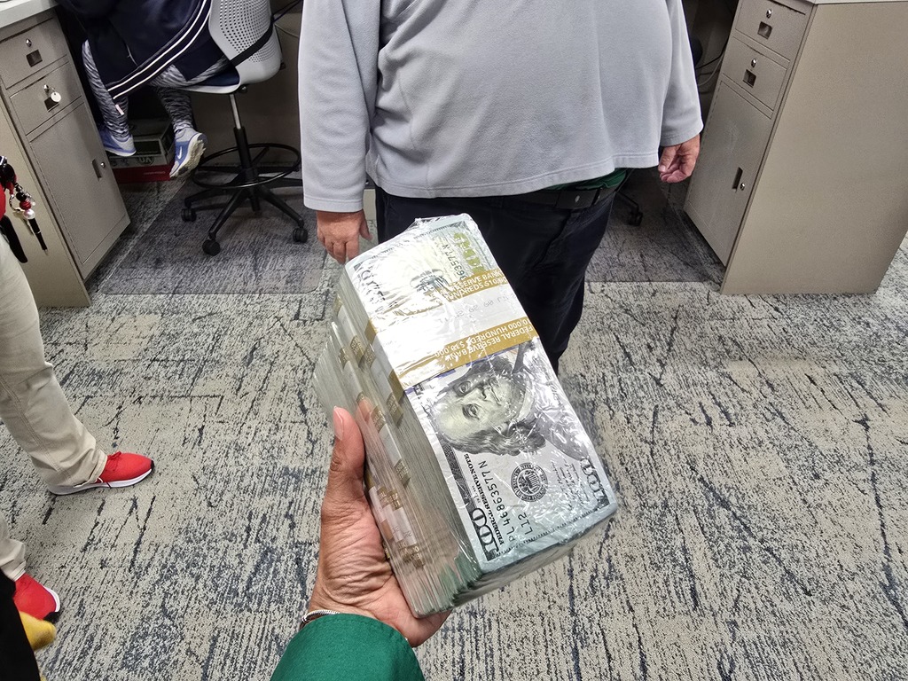 A person's hand holding a stack of cash, likely US dollars, on a carpeted office floor with filing cabinets in the background.