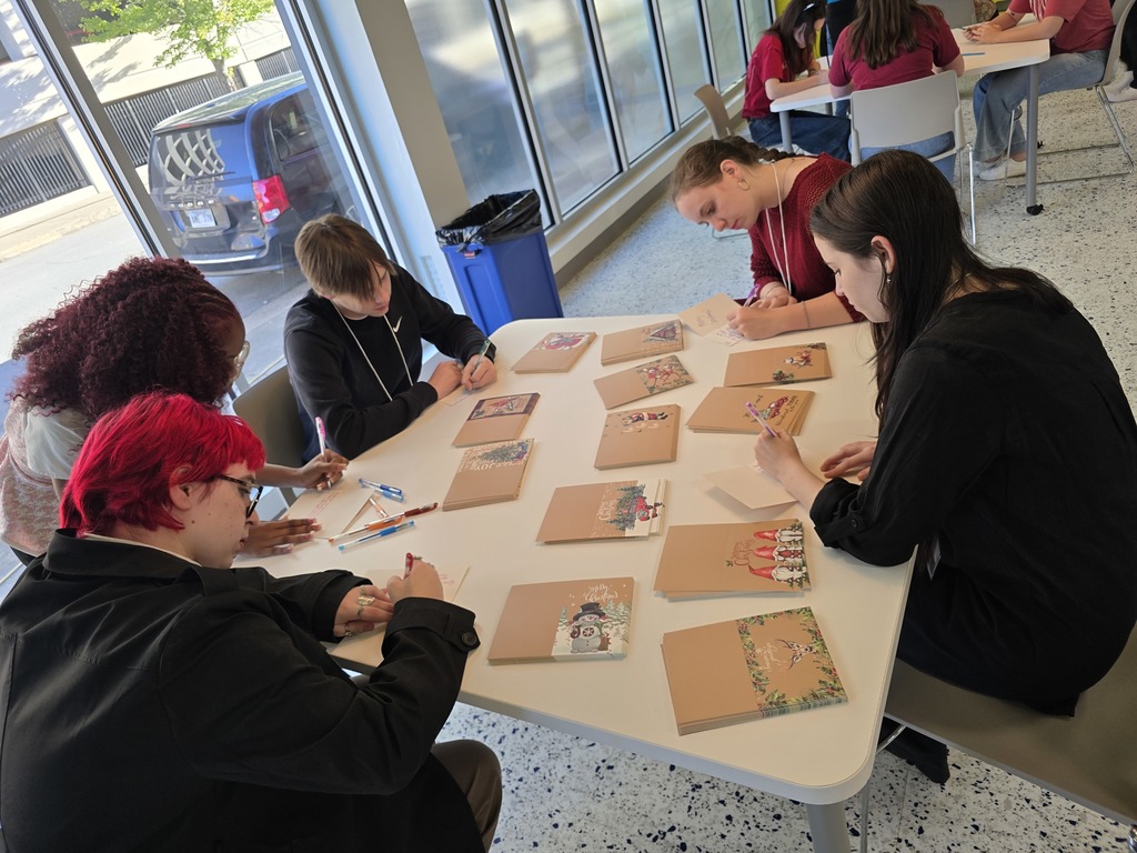 A group of people seated around a table, examining what appears to be playing cards or documents. The group appears to be engaged in a collaborative activity or discussion.