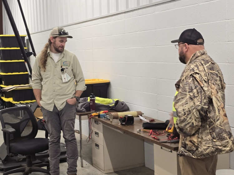 Two men in work attire conversing at a workbench with various tools and equipment.