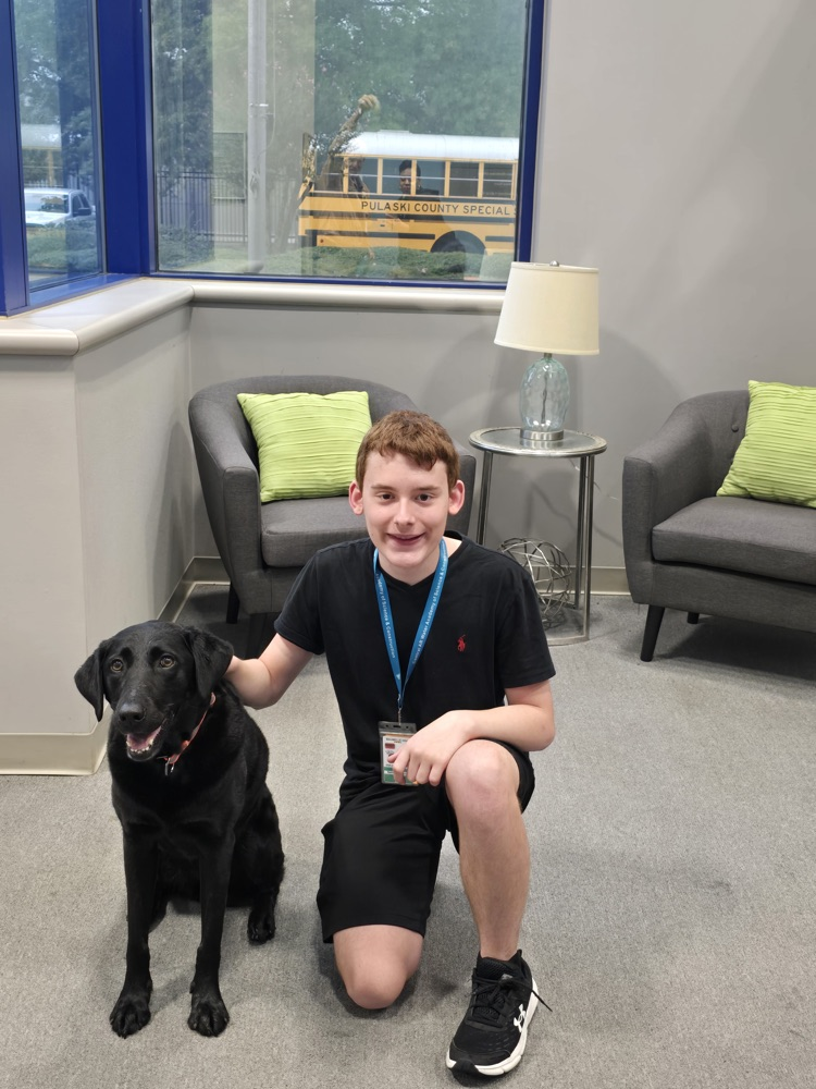 Young man in black shirt sitting next to black dog in a room with chairs and a school bus visible through the window.