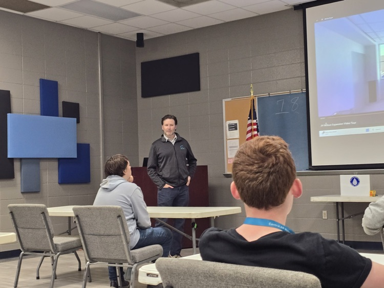 A group of people, including a man standing at the front, seated in a classroom-like setting with audio/visual equipment.