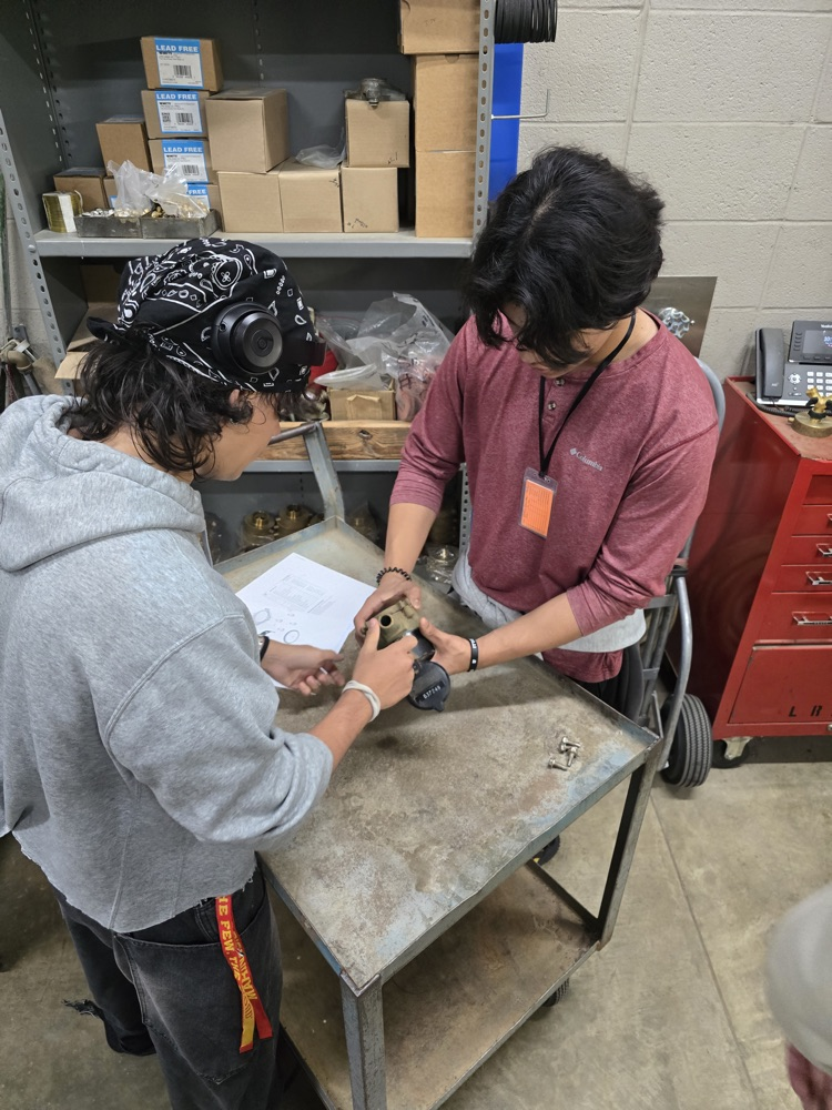 Two people working on computers in a workshop setting, surrounded by shelves and boxes.