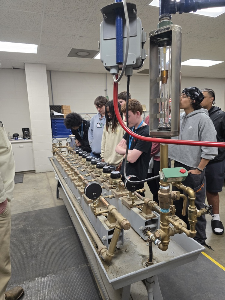 The image shows a group of people gathered around a large, complex machinery setup in what appears to be a laboratory or workshop setting. There are various equipment, tools, and components visible on the workbench.