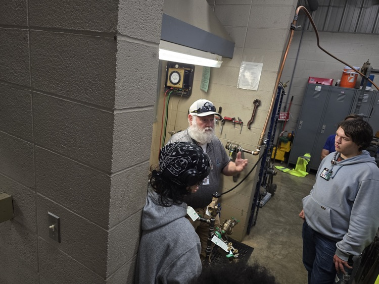 Two people in work uniforms in a utility room, with a clock on the wall and various tools and equipment visible.
