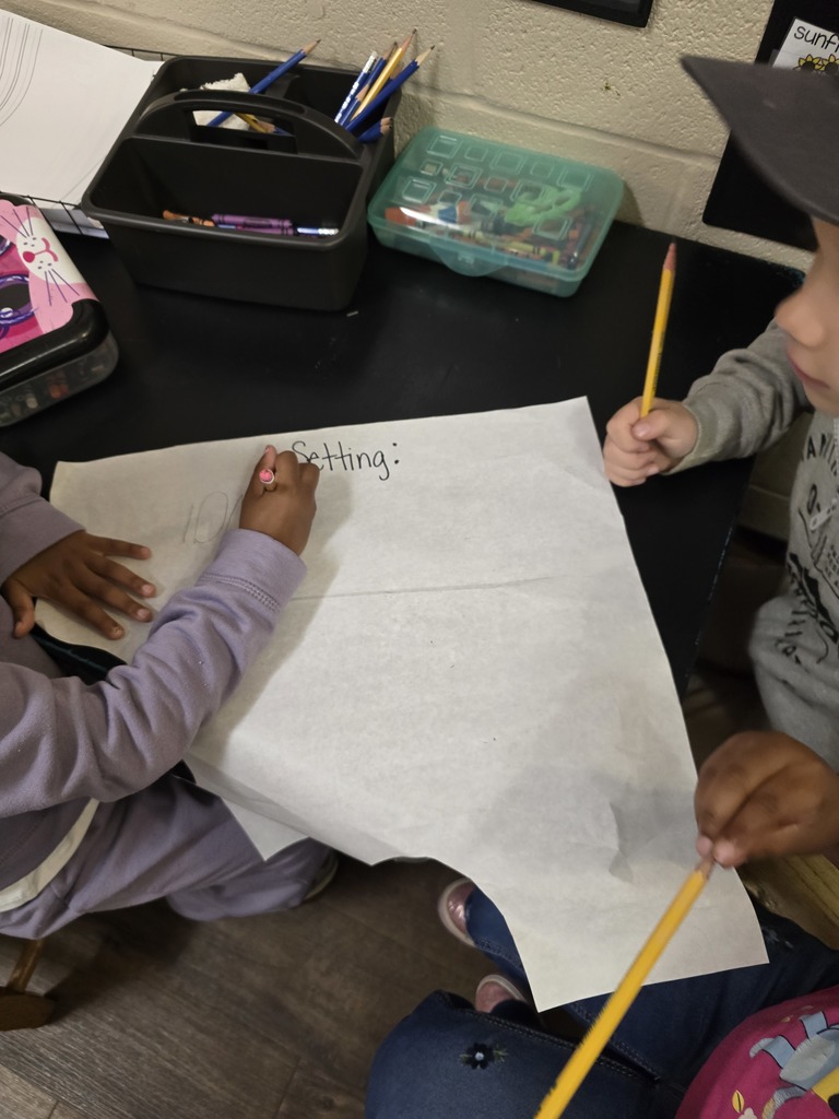 Two hands writing on paper in a school setting, with a desk, supplies, and other student materials visible.
