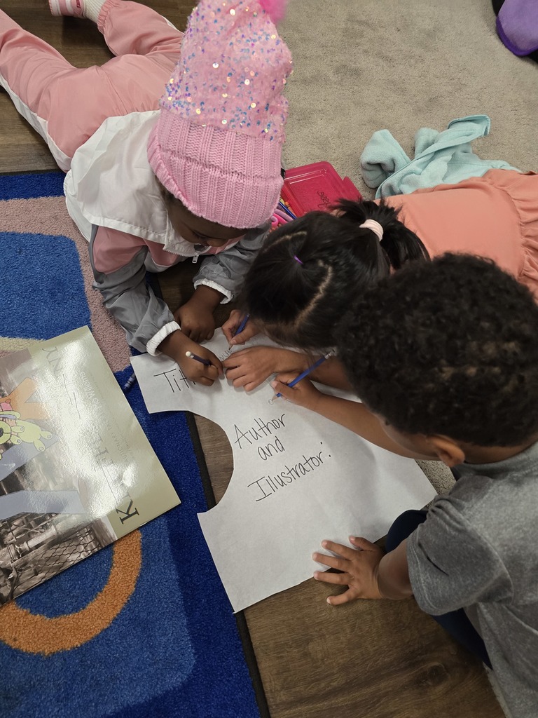 A person wearing a pink hat is working on a drawing on the floor surrounded by books and papers, and two students