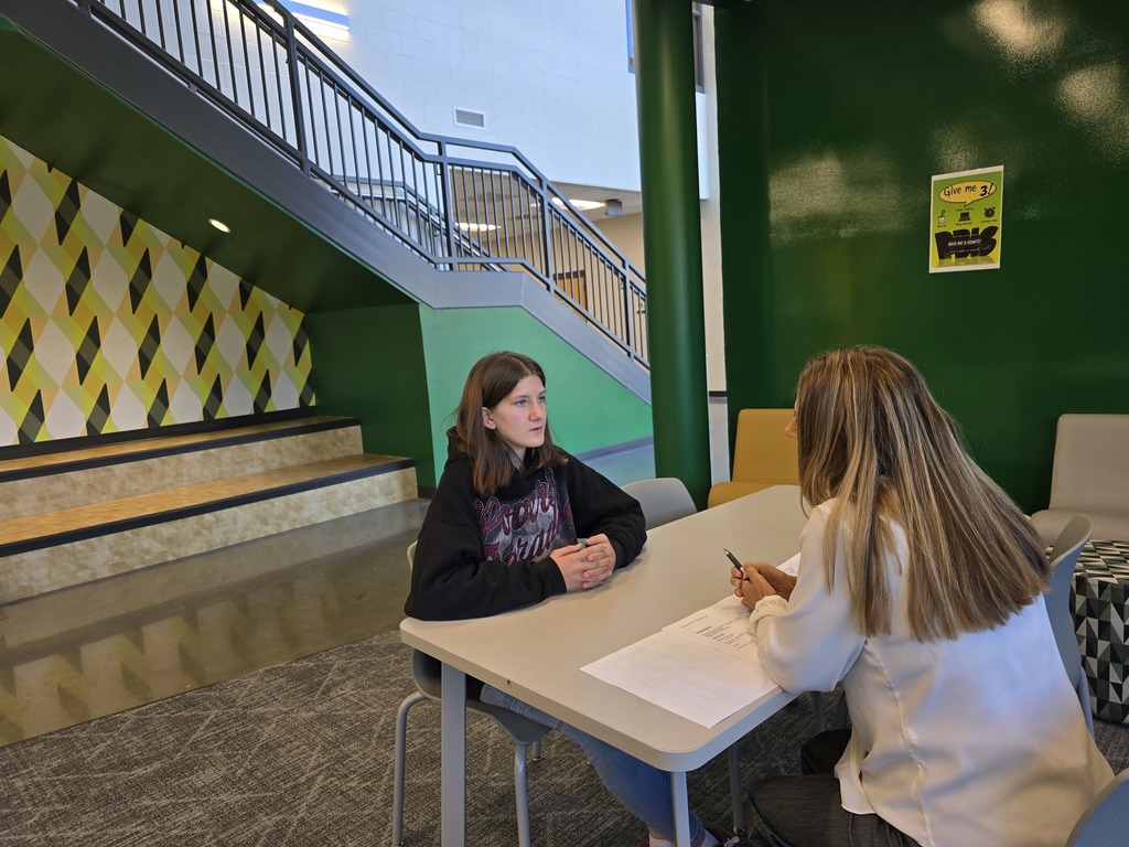 20251103_115223Two women sitting at a table in a building with multicolored walls and stairs. They are doing a mock interview.