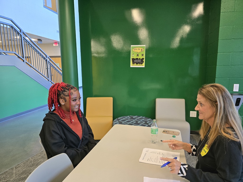 Two women sitting at a table in colorful room with green wall and signage. They are doing a mock interview.