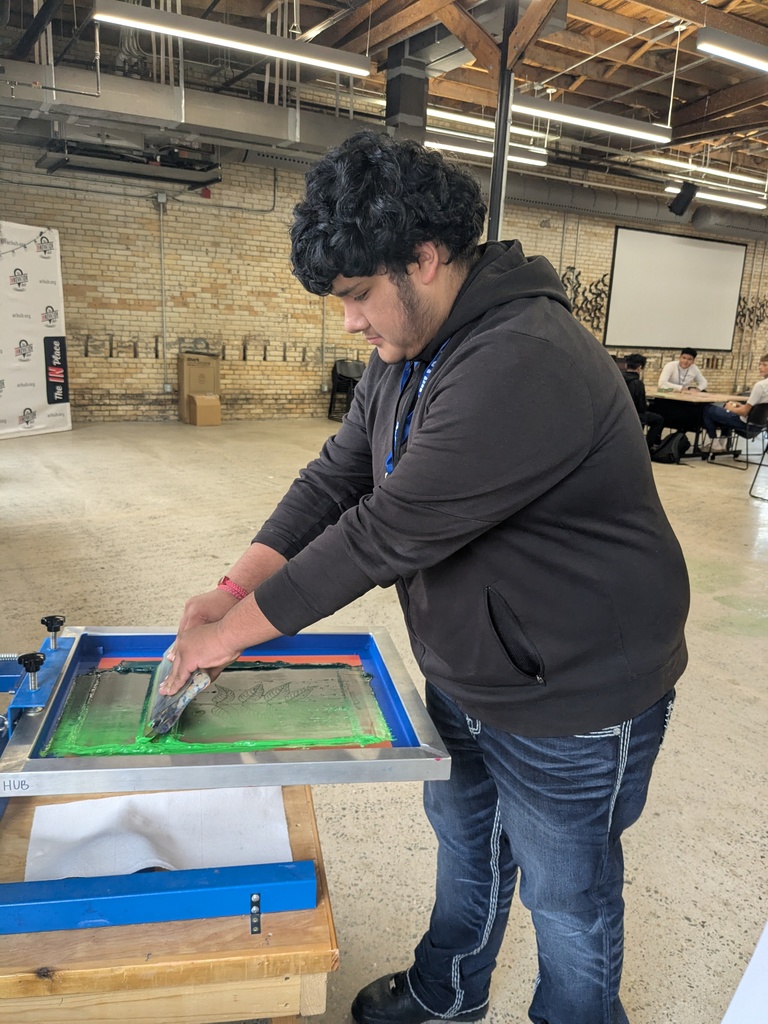 A person in a dark hoodie standing at a worktable, using a tool to work on a project in what appears to be a workshop or studio setting.