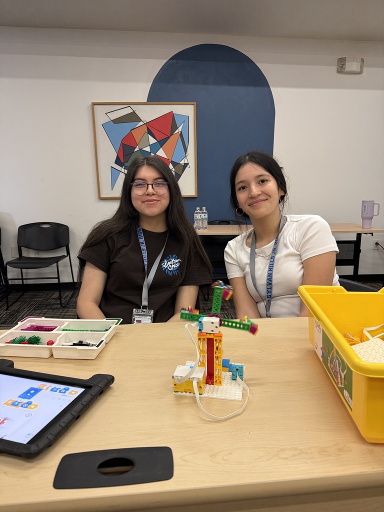 Two young women sitting at a table with electronic devices and craft materials in a classroom-like setting.
