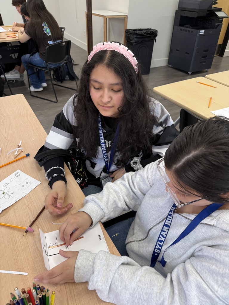 Two students working together at a desk, one wearing a pink headband and the other in a gray sweatshirt.