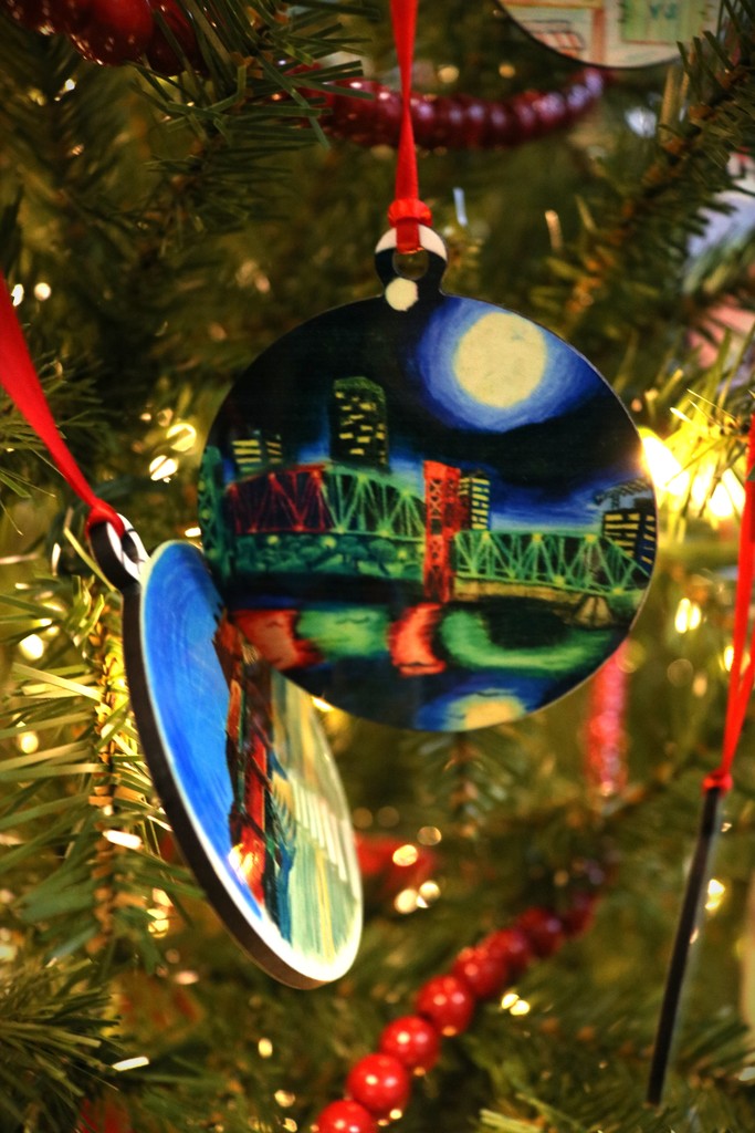 An ornament hanging from a Christmas tree. It shows an artist's rendition of a nighttime scene of a bridge over the Arkansas River with the Little Rock skyline.