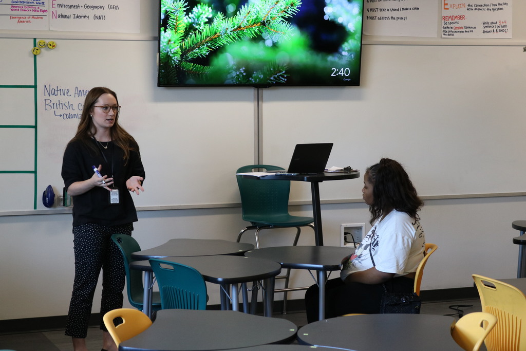 Two people in a classroom setting, one person standing and one seated at a desk, with a large display screen on the wall in the background.