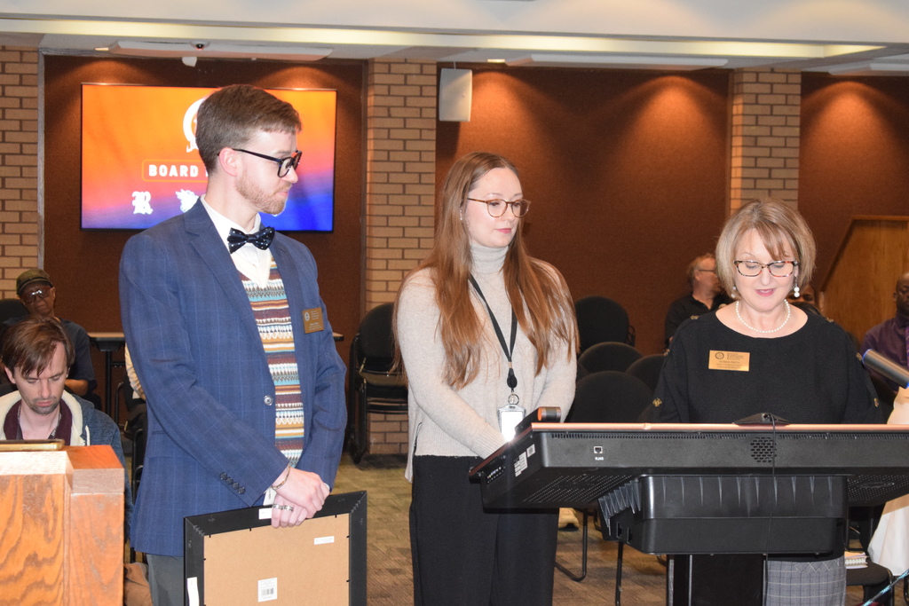 Three people standing at a podium, one woman in the center speaking into a microphone.