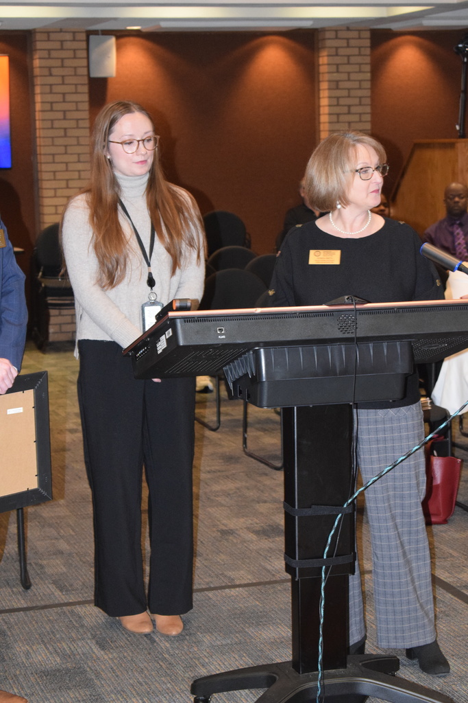 Two women at a podium, one younger with long hair and the other older with shorter hair, speaking at what appears to be an event or presentation.