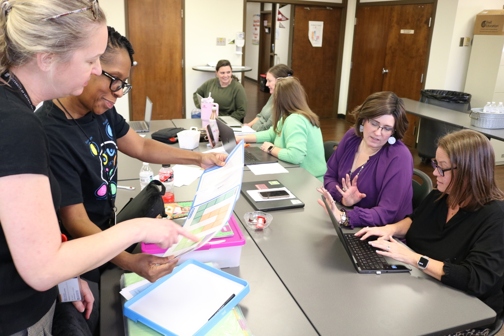 A group of people around a table, discussing documents and using laptop computers. The people are wearing a variety of casual and professional attire. There are also some office supplies on the table.