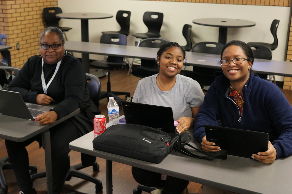 Three adults sitting together at a table, smiling and working on laptops in a classroom setting.