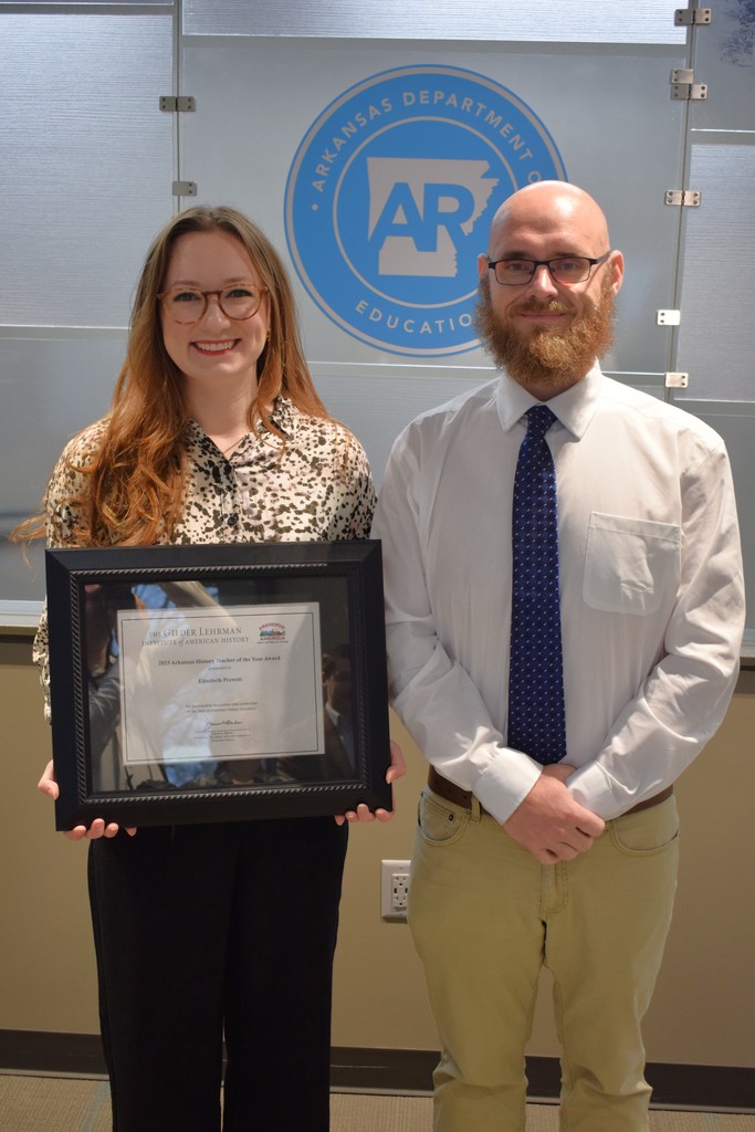 Two people standing in front of a sign for the Arkansas Department of Education, one holding a framed certificate.