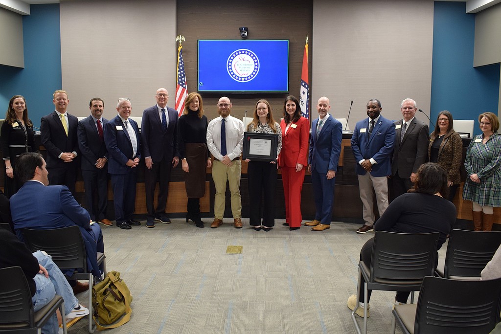 A group of people gathered for an ADE board meeting in front of a large flag, some seated and others standing.