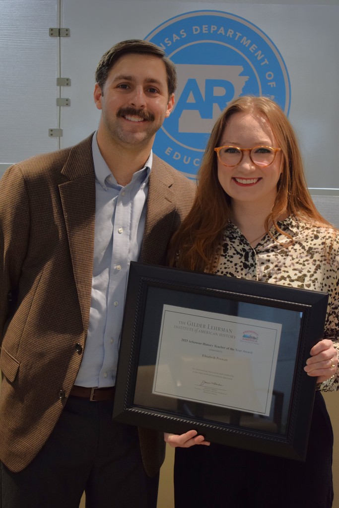A man and a woman smiling while holding an award certificate in front of a sign for the Arkansas Department of Education.