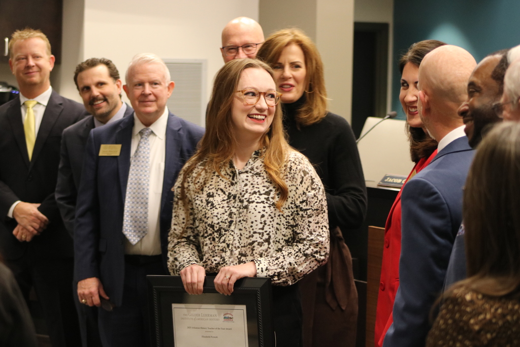 A large group of people in business attire gathered for what appears to be an awards ceremony or formal event, with a woman in the center smiling and holding an award or certificate.
